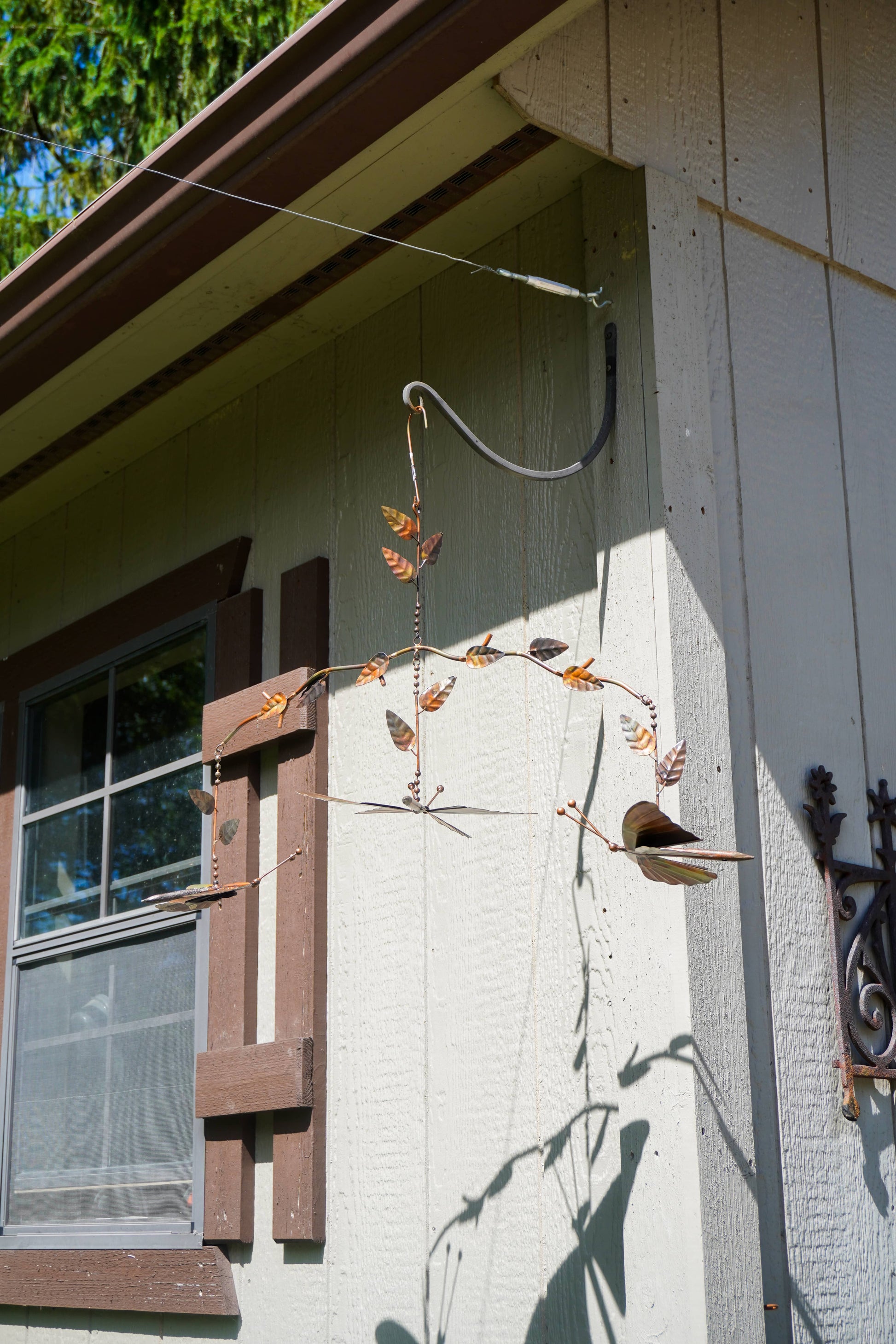 A rustic metal butterfly mobile hanging from a wire frame against the exterior wall of a house with beige siding and brown trim.