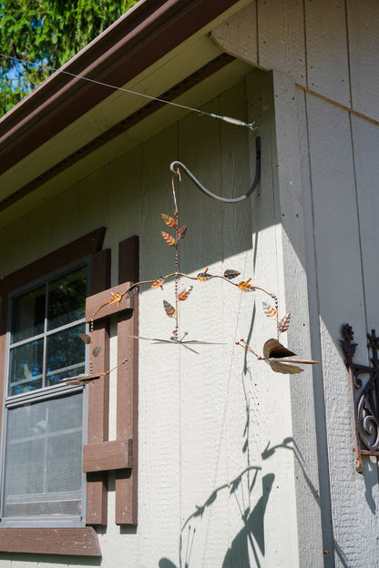 A rustic metal butterfly mobile hanging from a wire frame against the exterior wall of a house with beige siding and brown trim.