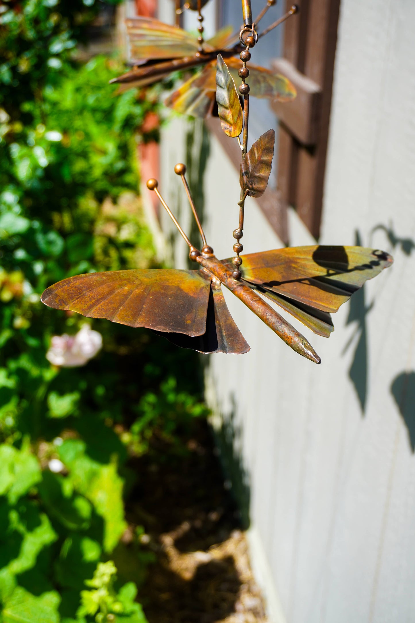 A metal butterfly garden sculpture in warm brown tones, hanging outdoors against a white wall with green foliage on the left.