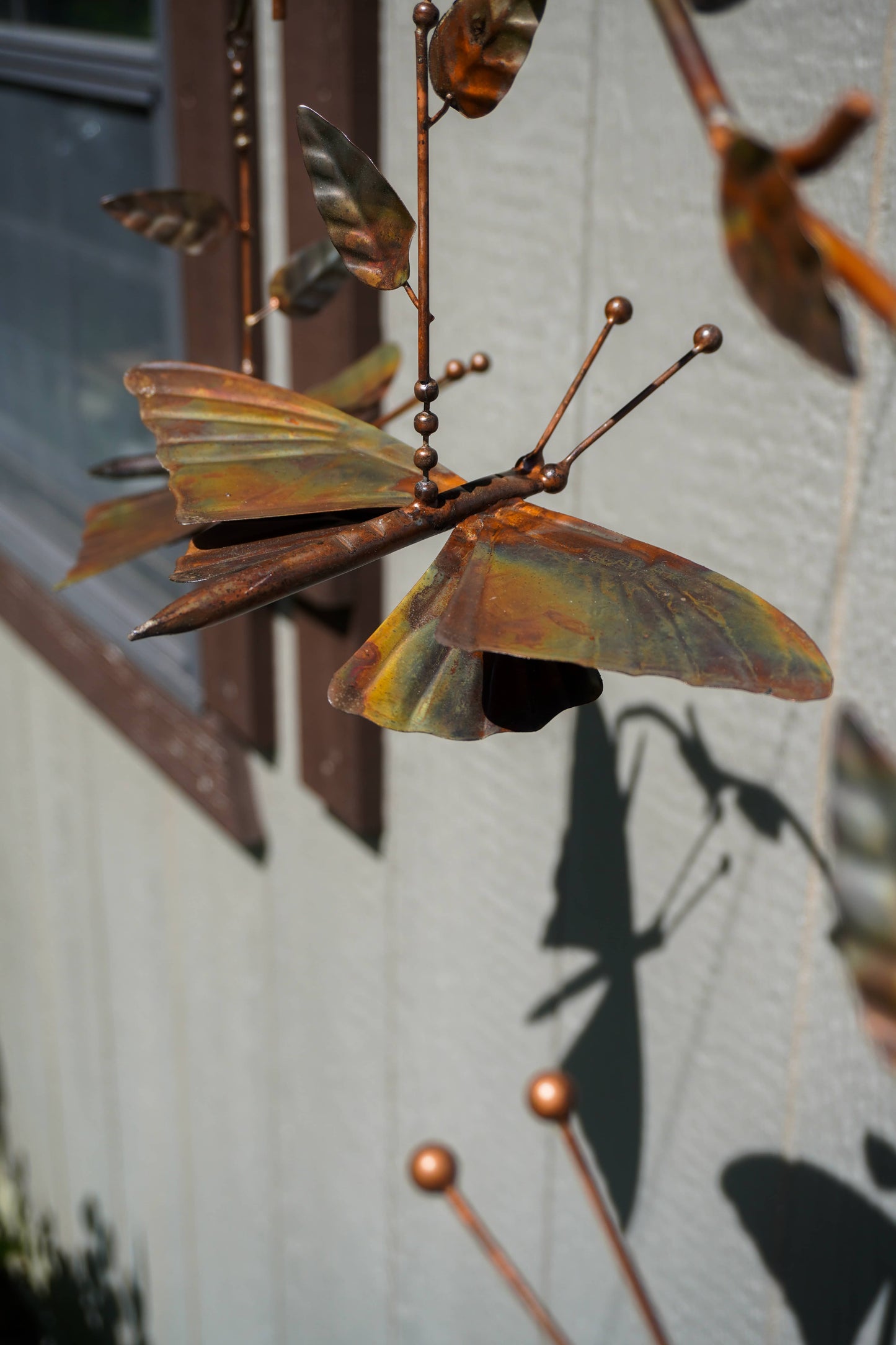 Metallic copper-colored butterfly garden sculpture hanging against a light gray stucco wall, with leaf-like wings and thin rod supports casting a shadow.