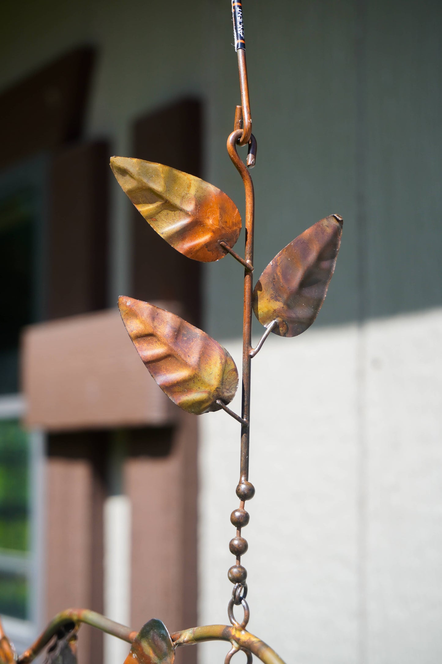 A close-up of a metal garden sculpture featuring three leaf-shaped elements in a distressed copper and brown finish, mounted on a vertical metal stem with bead accents.