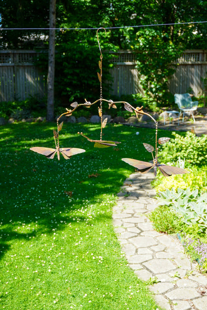 A metal butterfly garden mobile hanging outdoors in a backyard garden with green grass, a stone path, and trees in the background.