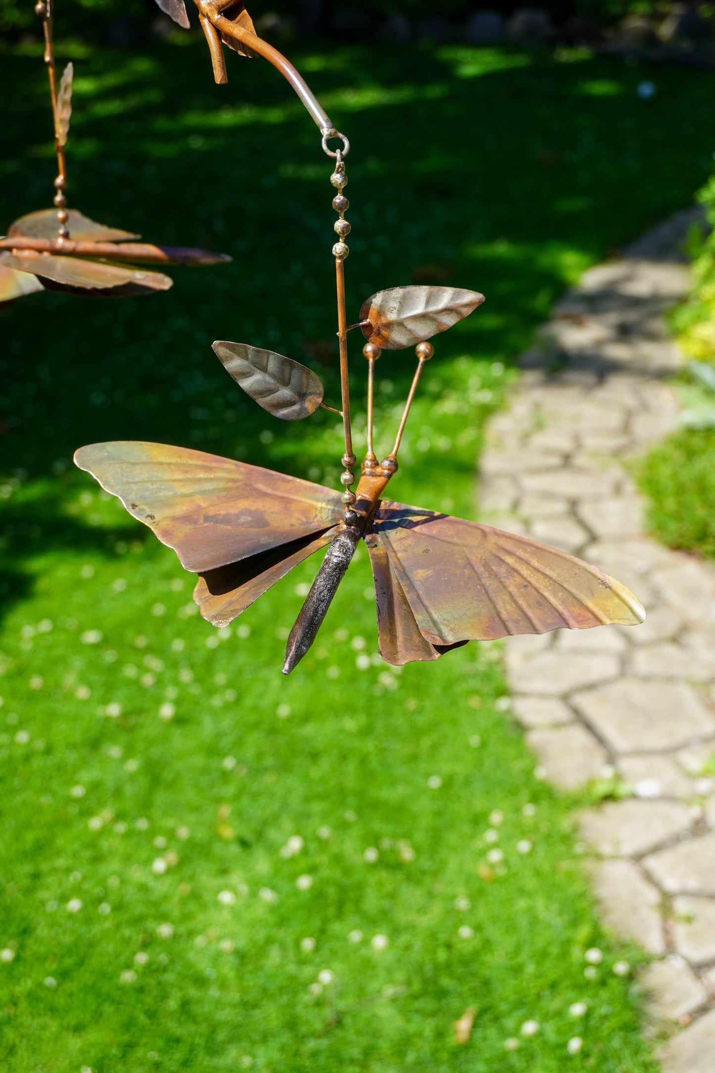 A bronze metal garden wind spinner featuring butterfly and leaf shapes hanging from a chain in an outdoor garden setting with green grass and a stone path.
