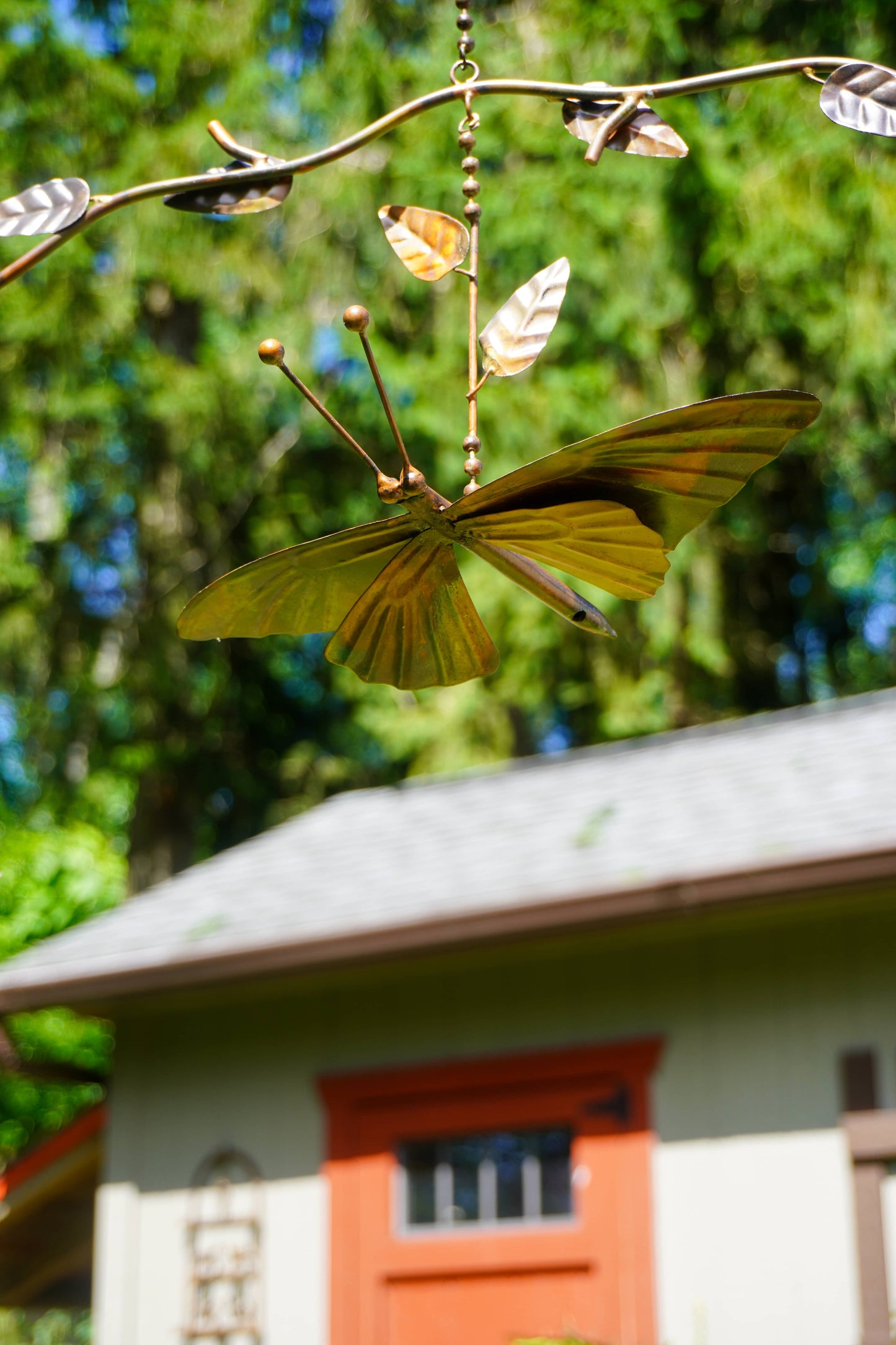 A metal garden wind spinner featuring a large butterfly and smaller leaves attached to a branch-shaped frame, photographed outdoors in front of a blurred house and trees.