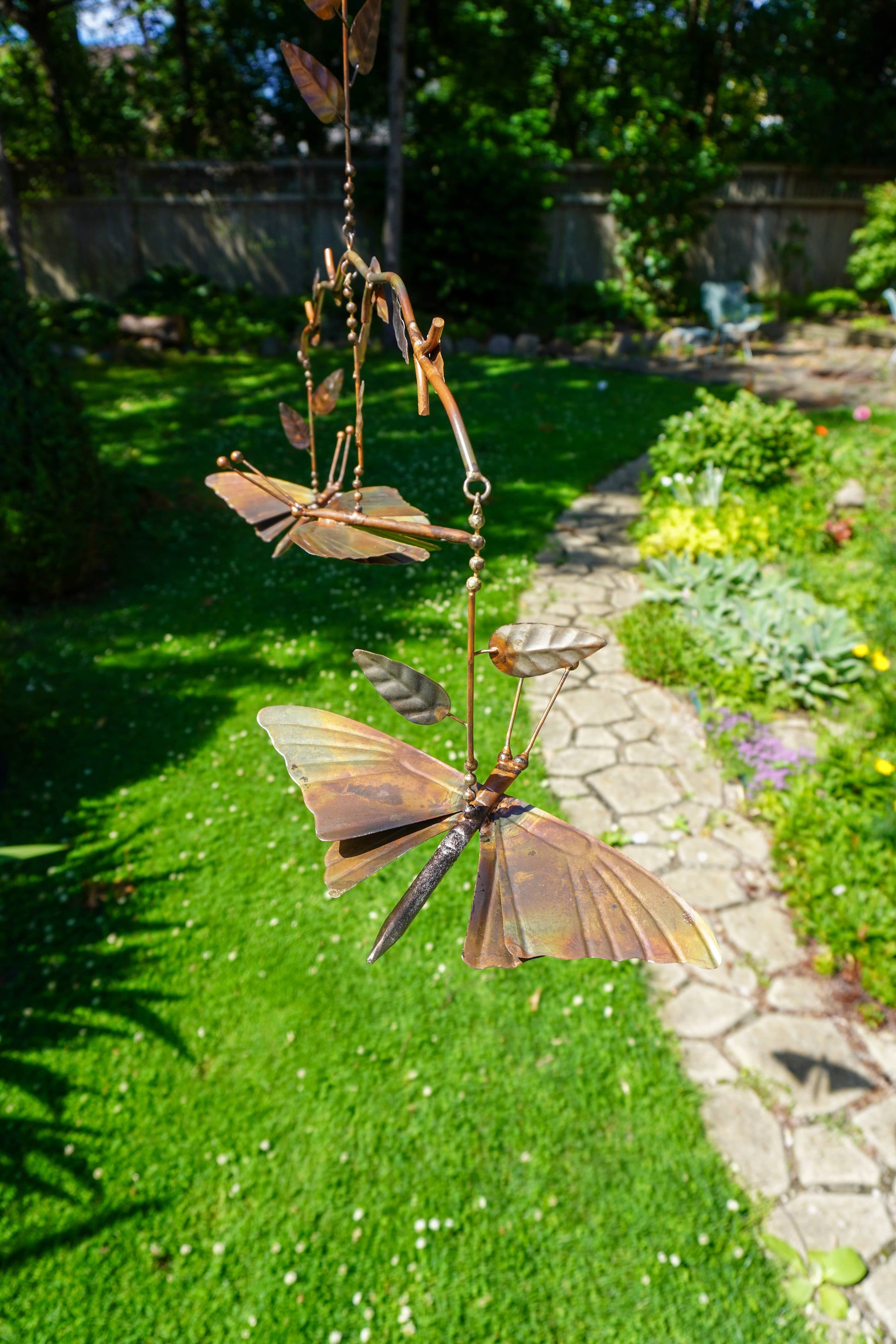 Metal butterfly garden wind spinner hanging in a sunny backyard garden with a stone path and green lawn.