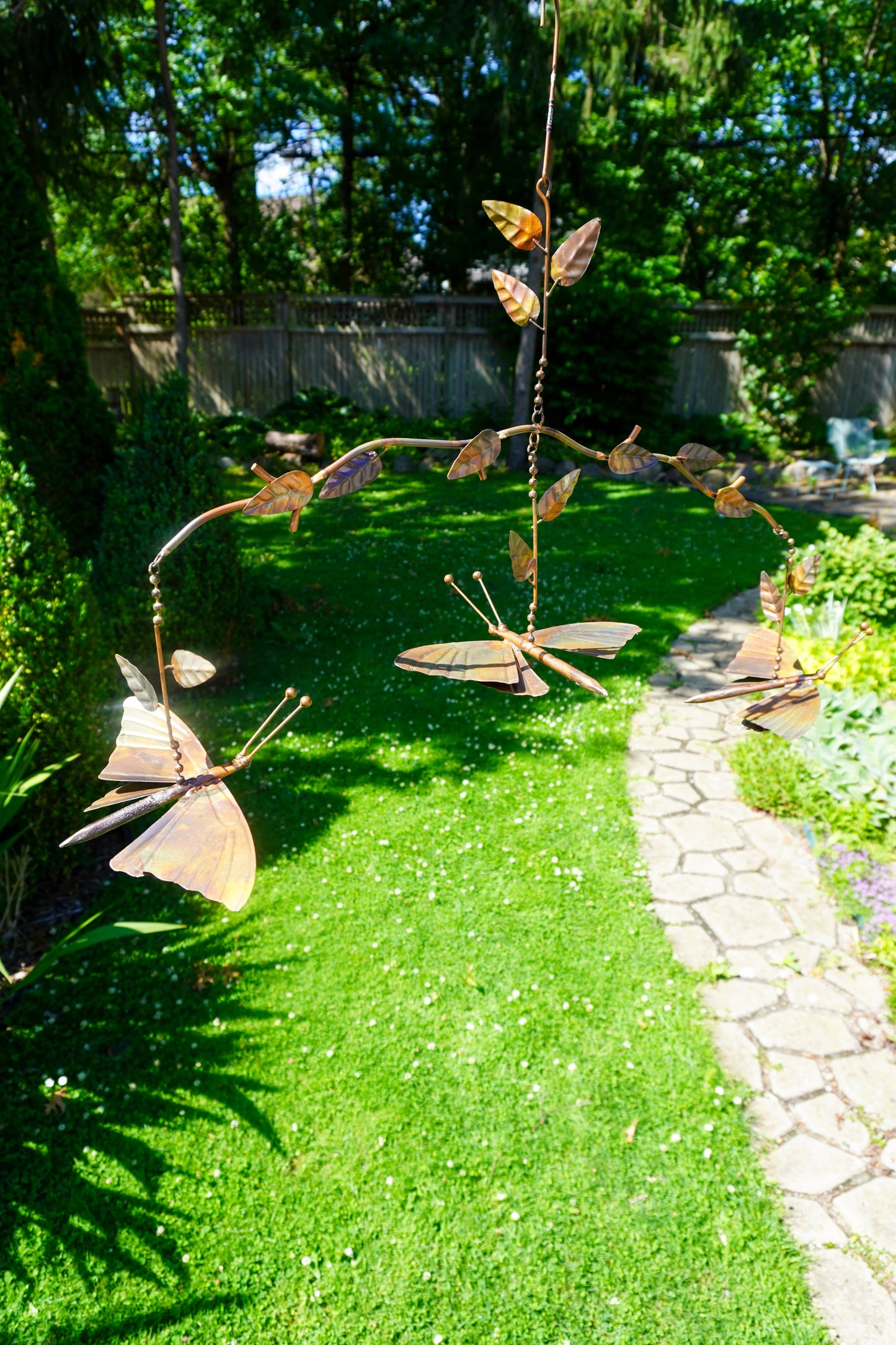 A metal garden mobile with butterfly shapes hangs over a green lawn beside a stone pathway in a backyard garden.