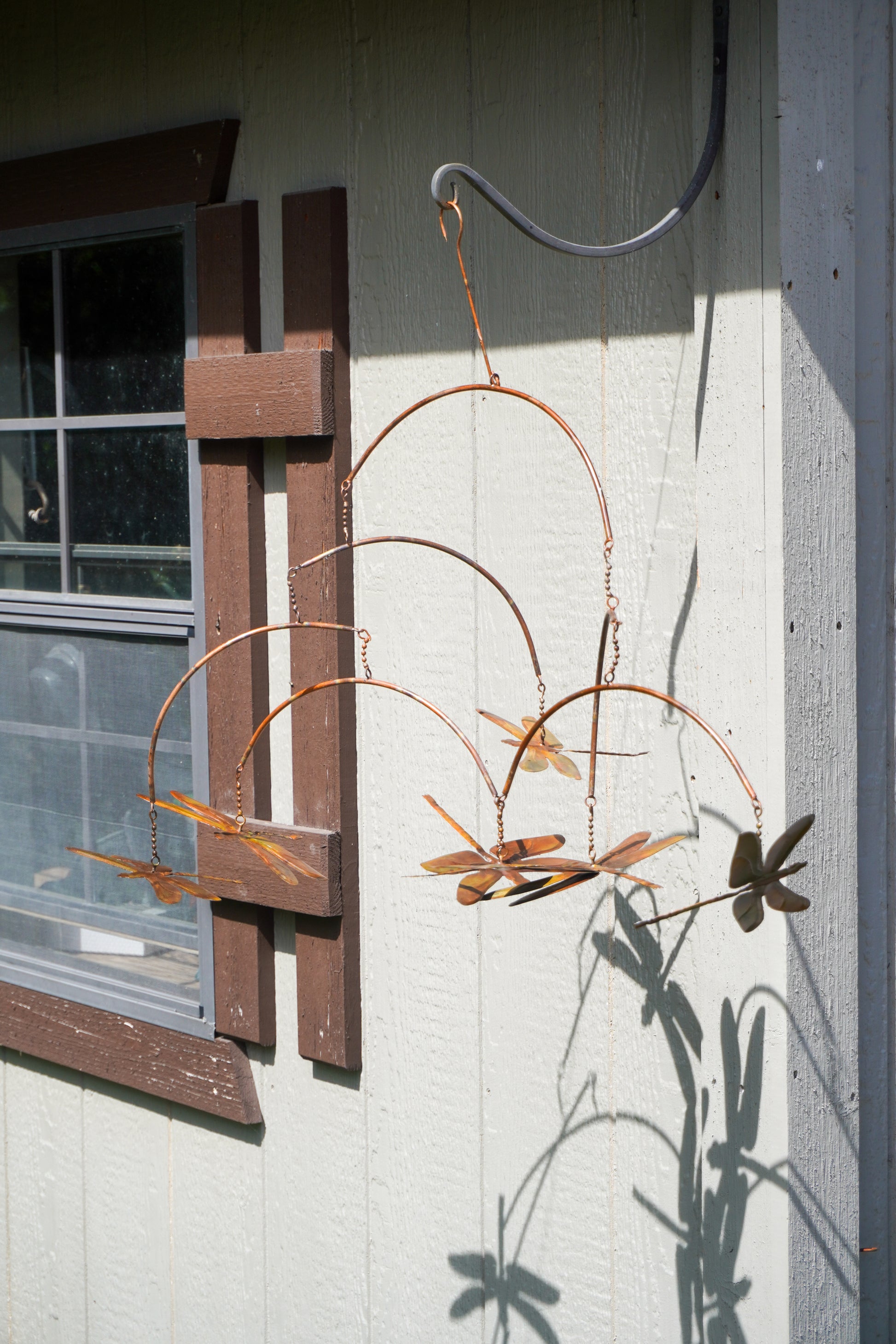A copper-colored metal dragonfly mobile hanging from a hook on a light-colored exterior wall next to a window.