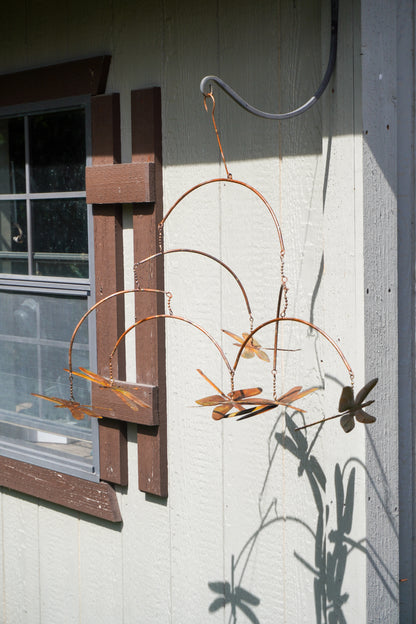 A copper-colored metal dragonfly mobile hanging from a hook on a light-colored exterior wall next to a window.