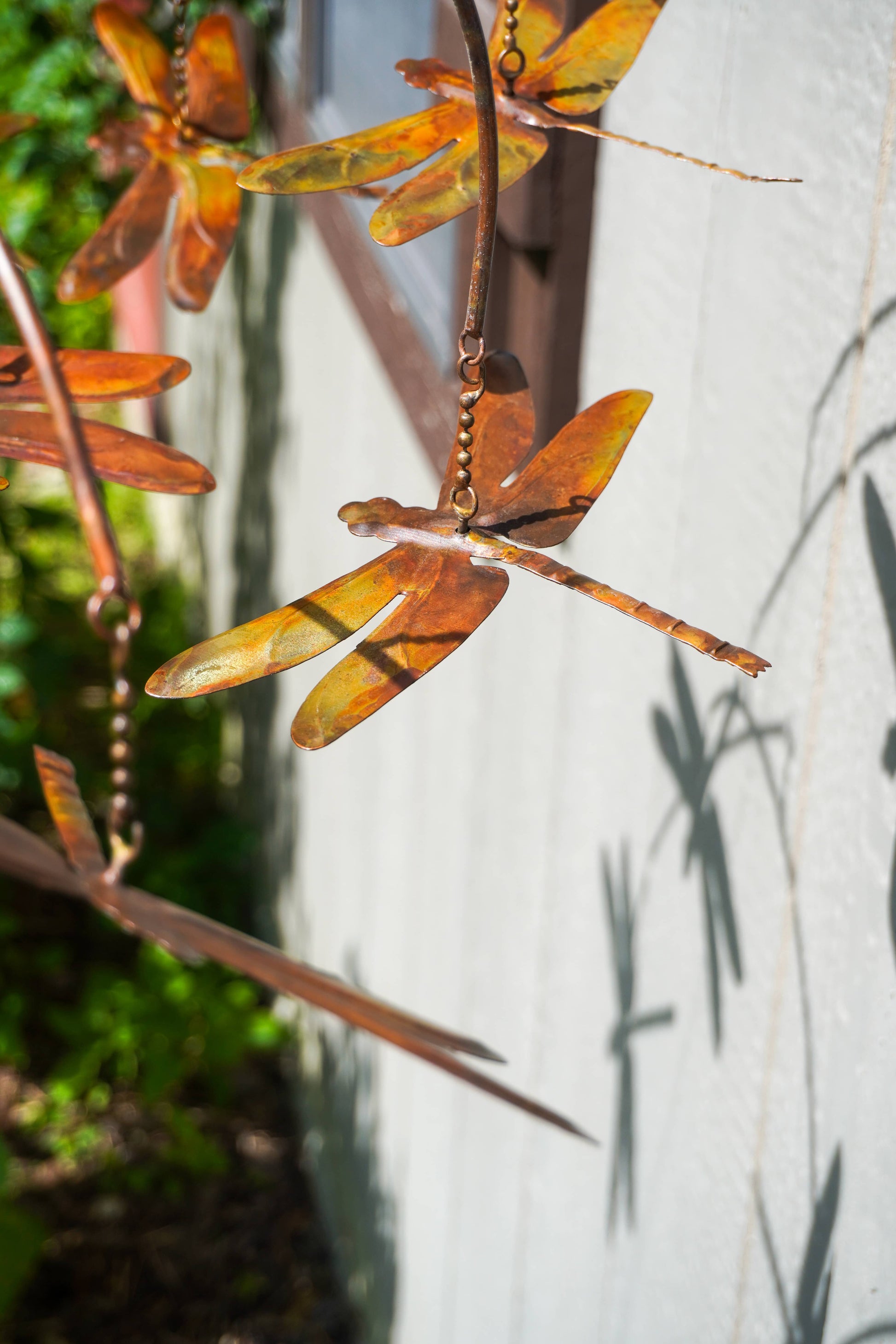 Metal dragonfly garden hanging sculpture in rusted orange-brown and bronze tones, shown outdoors against a white wall with leaf shadows.