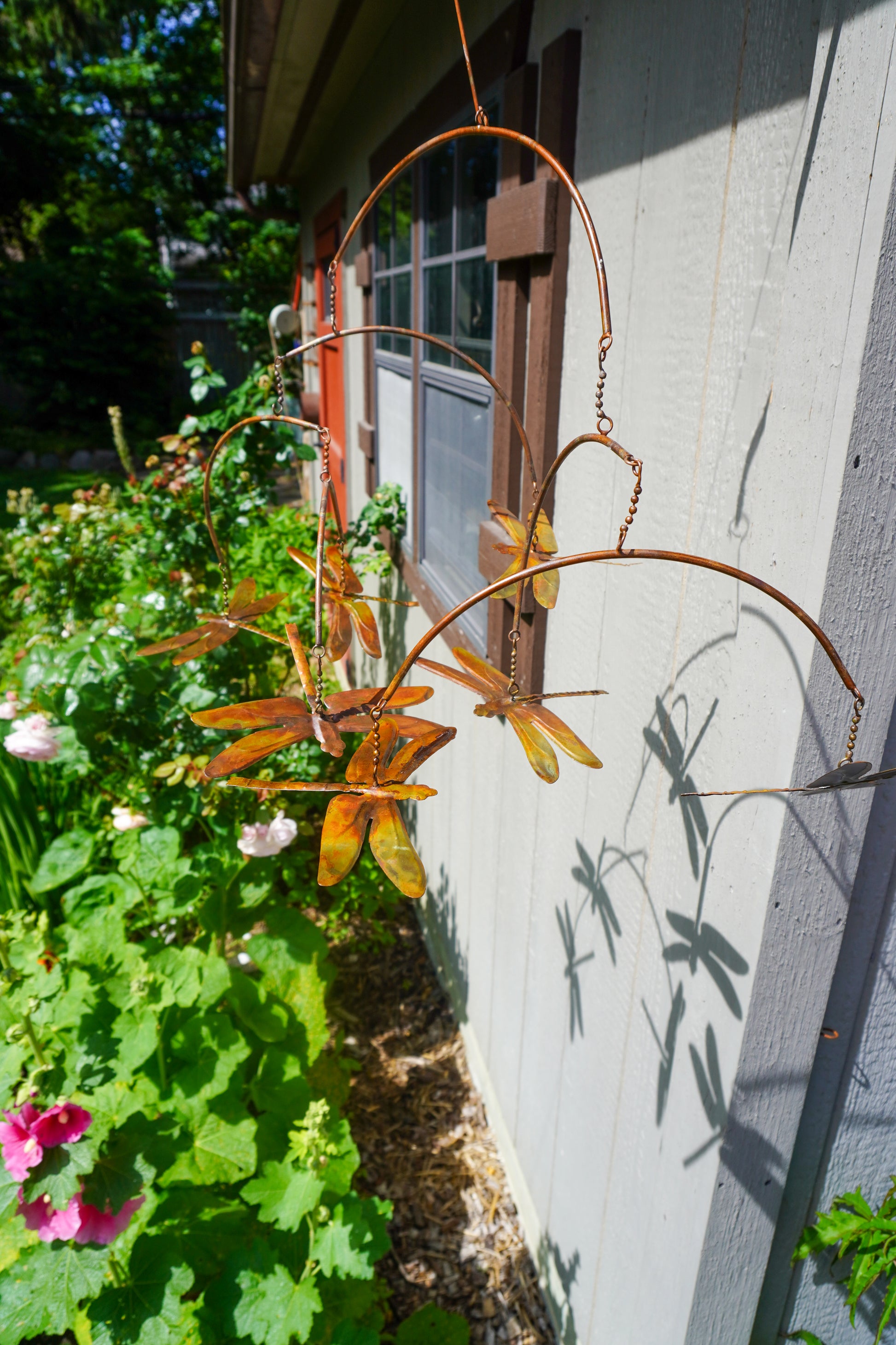 A copper-colored metal dragonfly garden hanging ornament suspended outdoors next to a house wall with green garden plants and flowers.