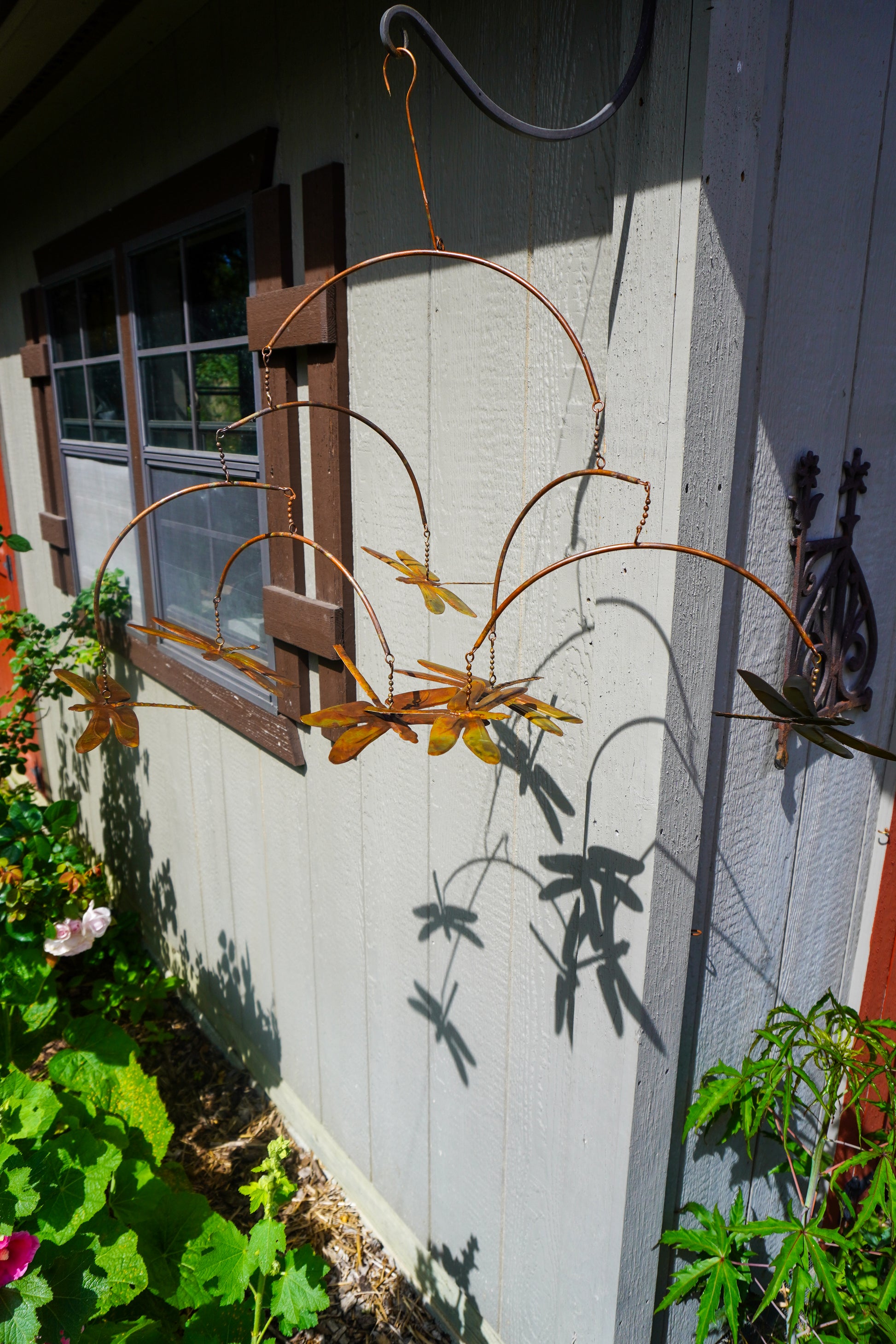 A rustic metal dragonfly garden swing hanging outside a house, casting a long shadow on the white siding. The swing features multiple curved metal arms and several small dragonfly cutouts in brown and yellow tones.