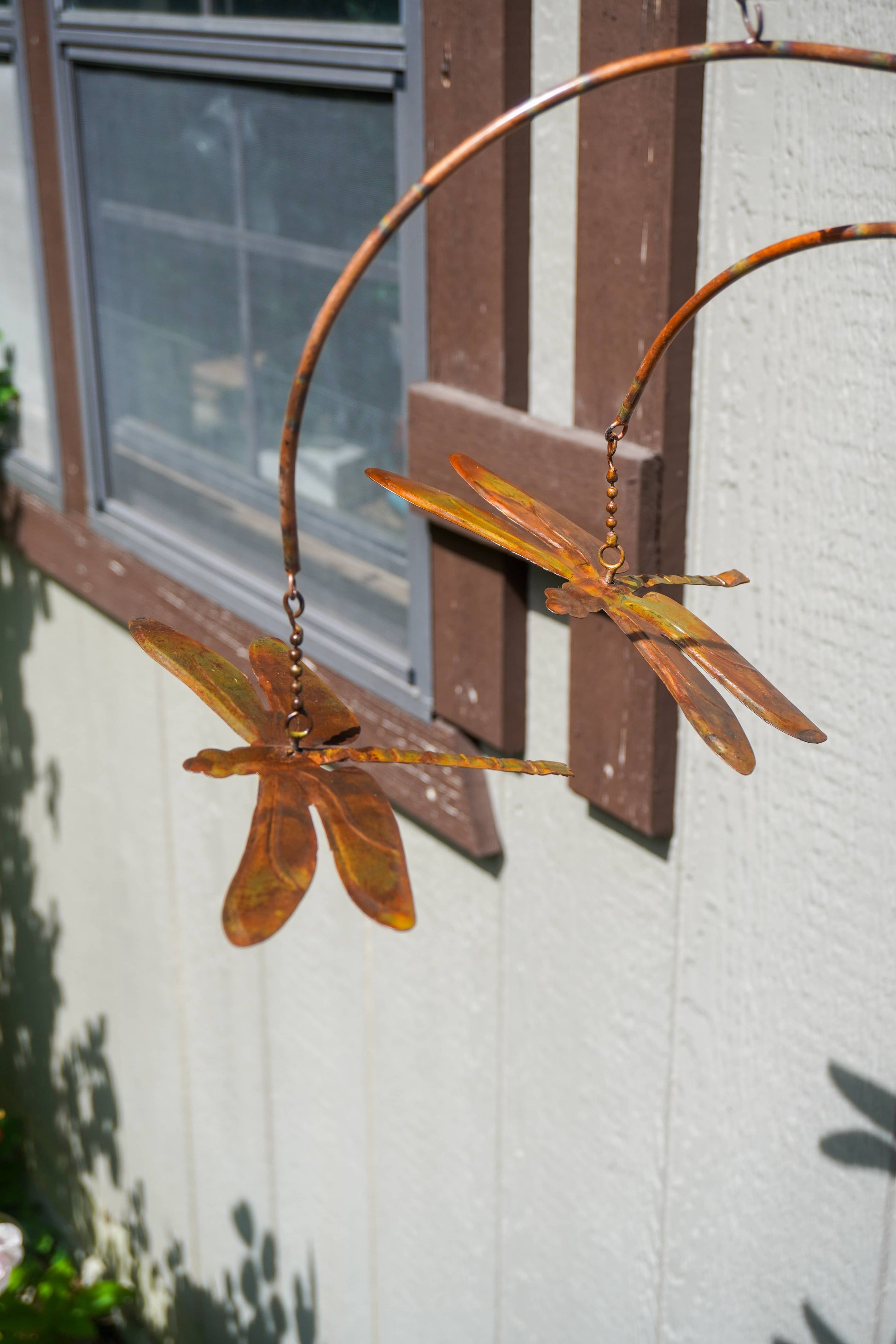 Two copper dragonfly garden sculptures hanging from a curved metal frame against an exterior house wall with a window.