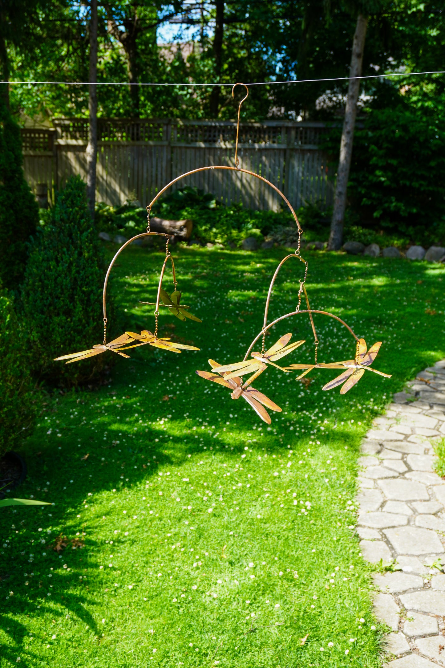 Metal dragonfly garden swings hanging from a wire in a sunny backyard lawn with green grass, a stone path, and trees in the background.