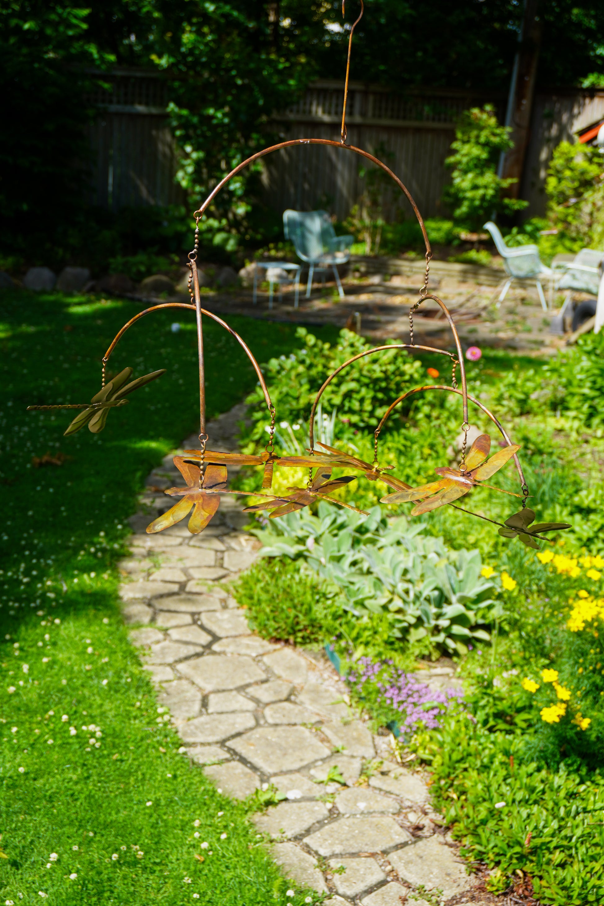 A metal garden swing with dragonfly motifs hanging in a lush green backyard garden next to a stone path.