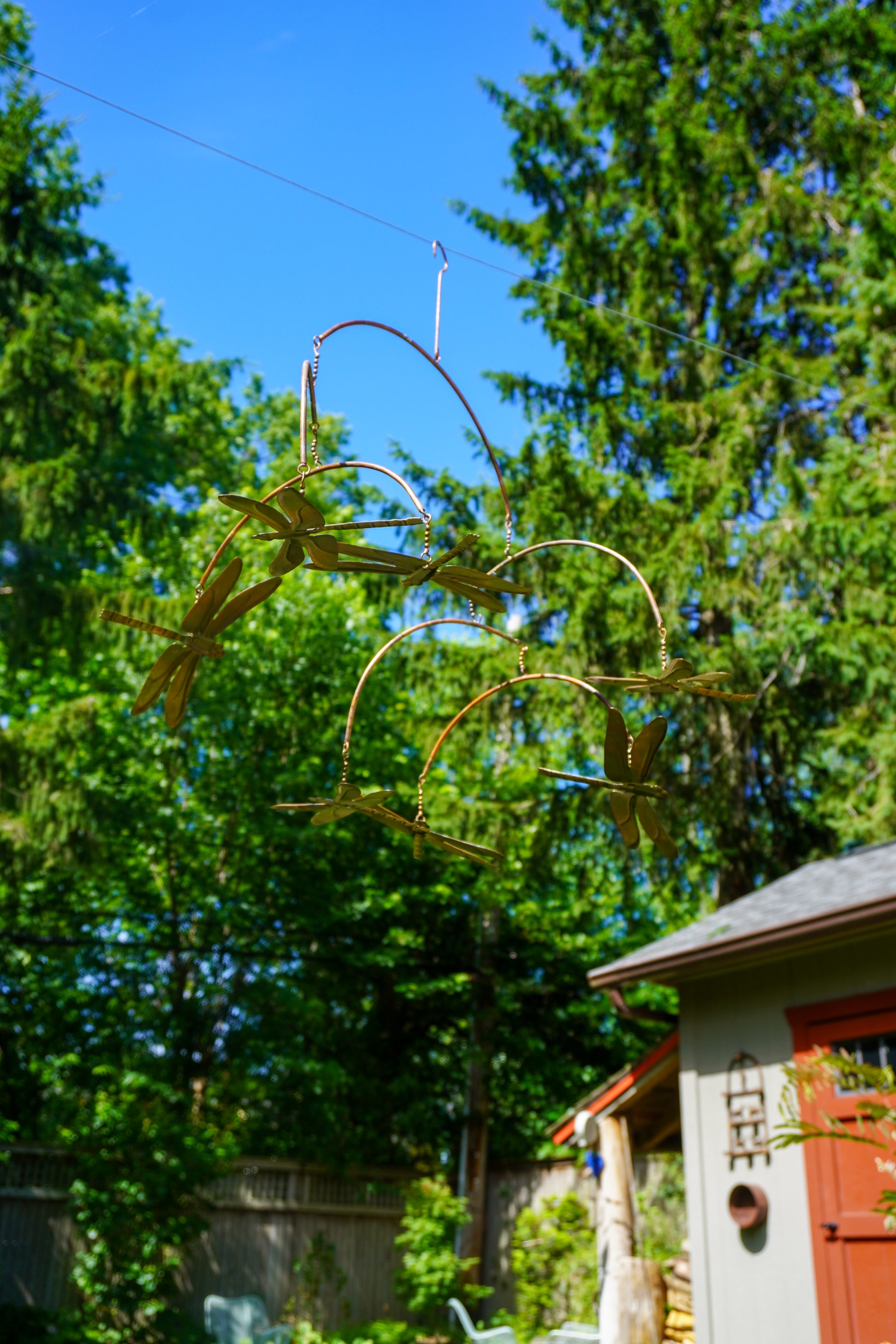 A metal hanging mobile with dragonfly wings suspended in a backyard with green trees, blue sky, and part of a shed visible.