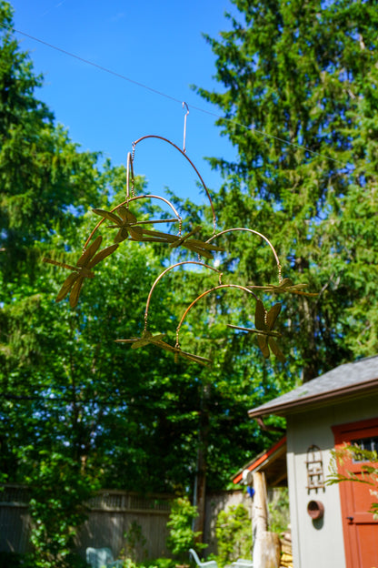 A metal hanging mobile with dragonfly wings suspended in a backyard with green trees, blue sky, and part of a shed visible.