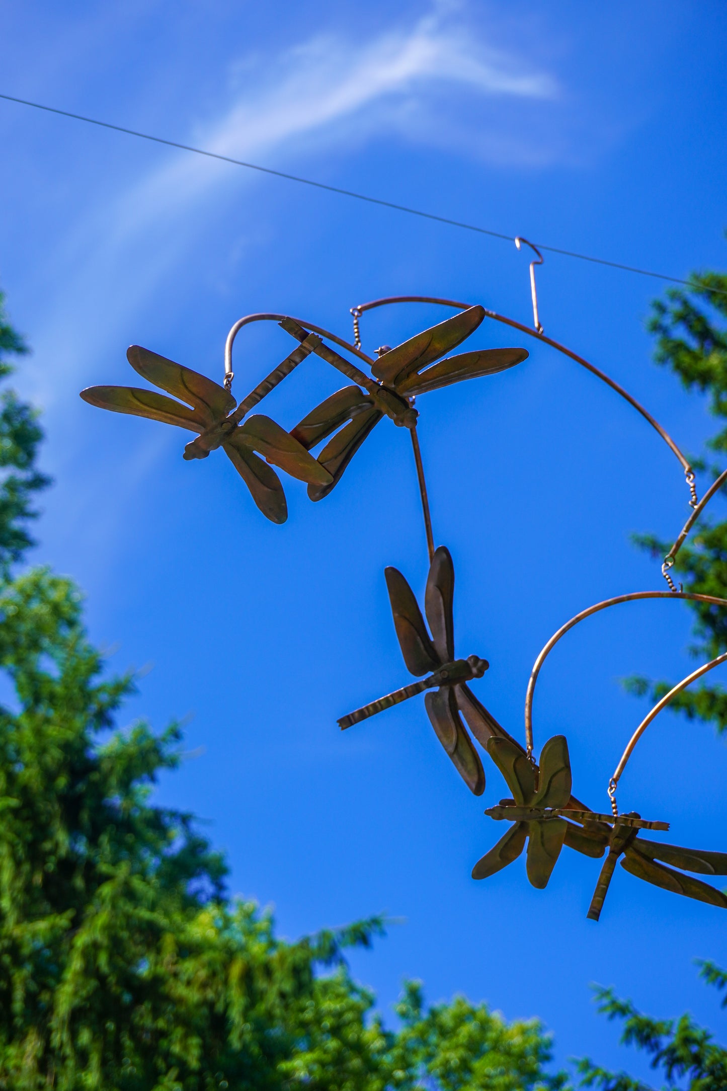 Metal dragonfly garden sculpture hanging outdoors against a bright blue sky with green tree foliage visible at the edges.