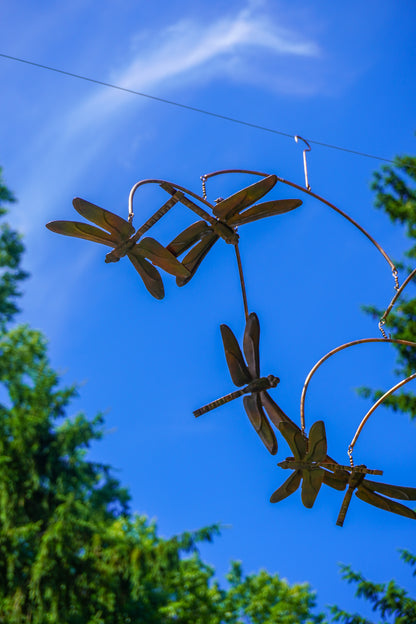 Metal dragonfly garden sculpture hanging outdoors against a bright blue sky with green tree foliage visible at the edges.