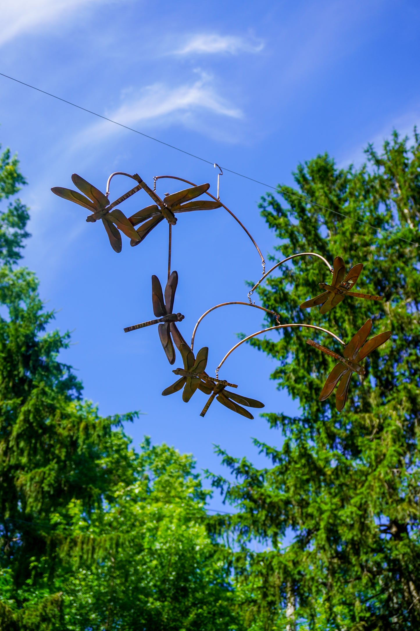 Metal dragonfly garden sculpture hanging outdoors against a bright blue sky and green trees.