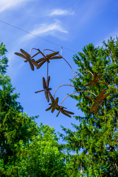 Metal dragonfly garden sculpture hanging outdoors against a bright blue sky and green trees.
