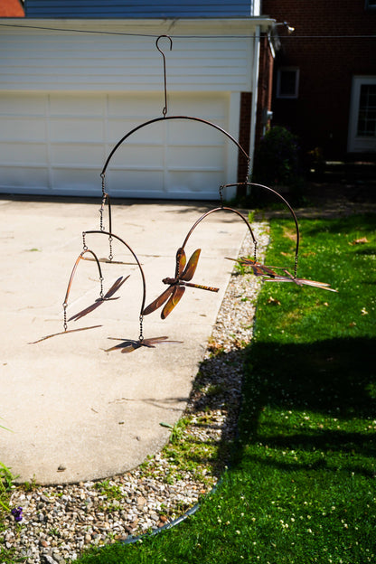 A rustic metal dragonfly hanging garden ornament suspended from a curved metal frame outdoors in a backyard setting with a concrete driveway, grass, and a garage in the background.