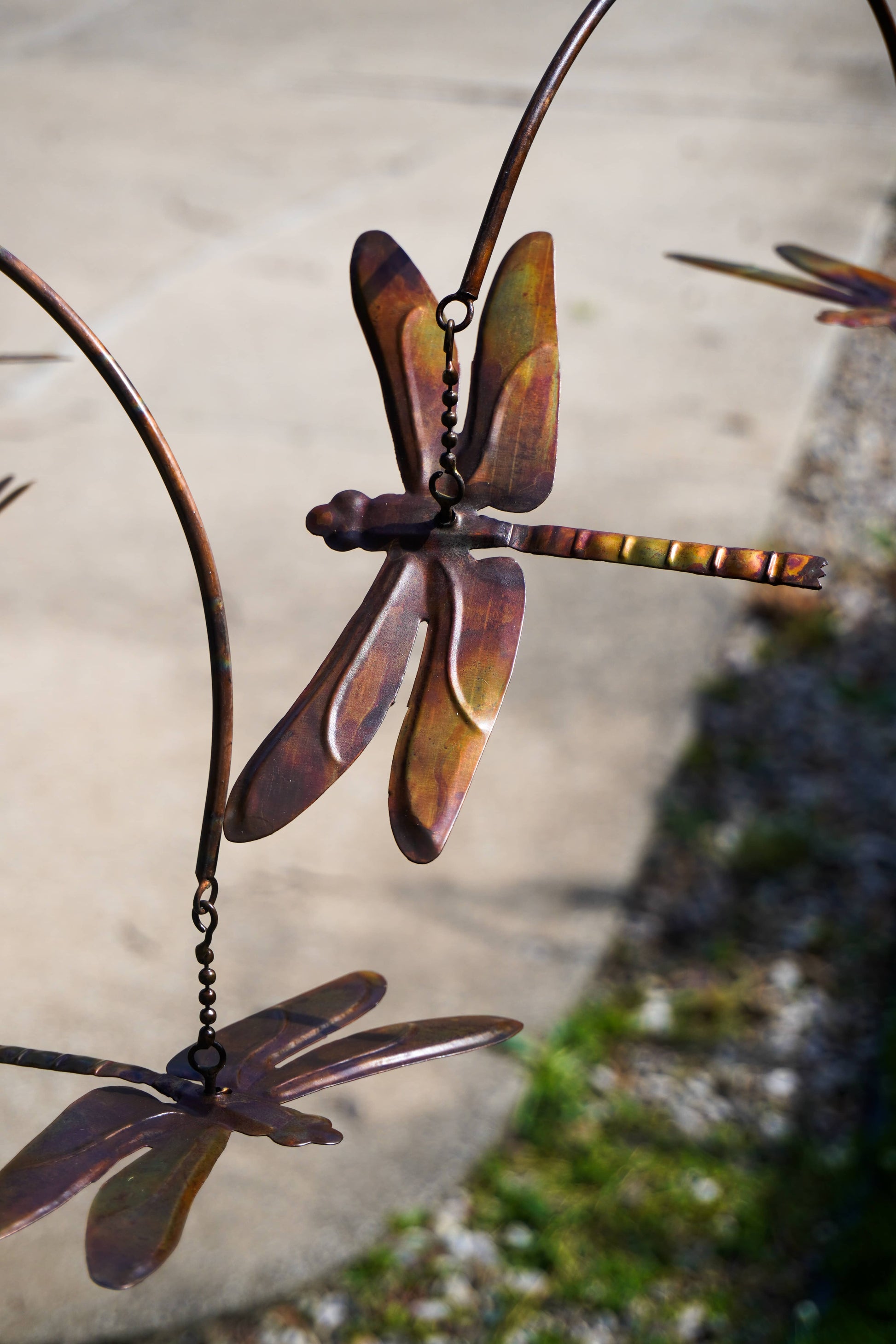 Metal dragonfly garden wind spinner with a rustic copper-brown finish, shown hanging outdoors against a blurred concrete and grass background.