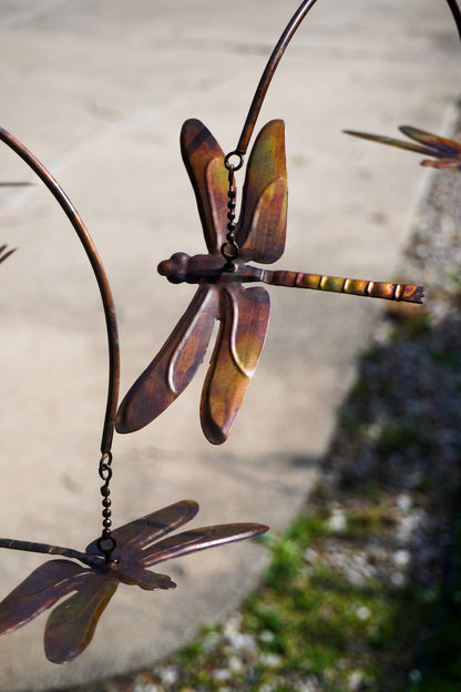 Metal dragonfly garden wind spinner with a rustic copper-brown finish, shown hanging outdoors against a blurred concrete and grass background.