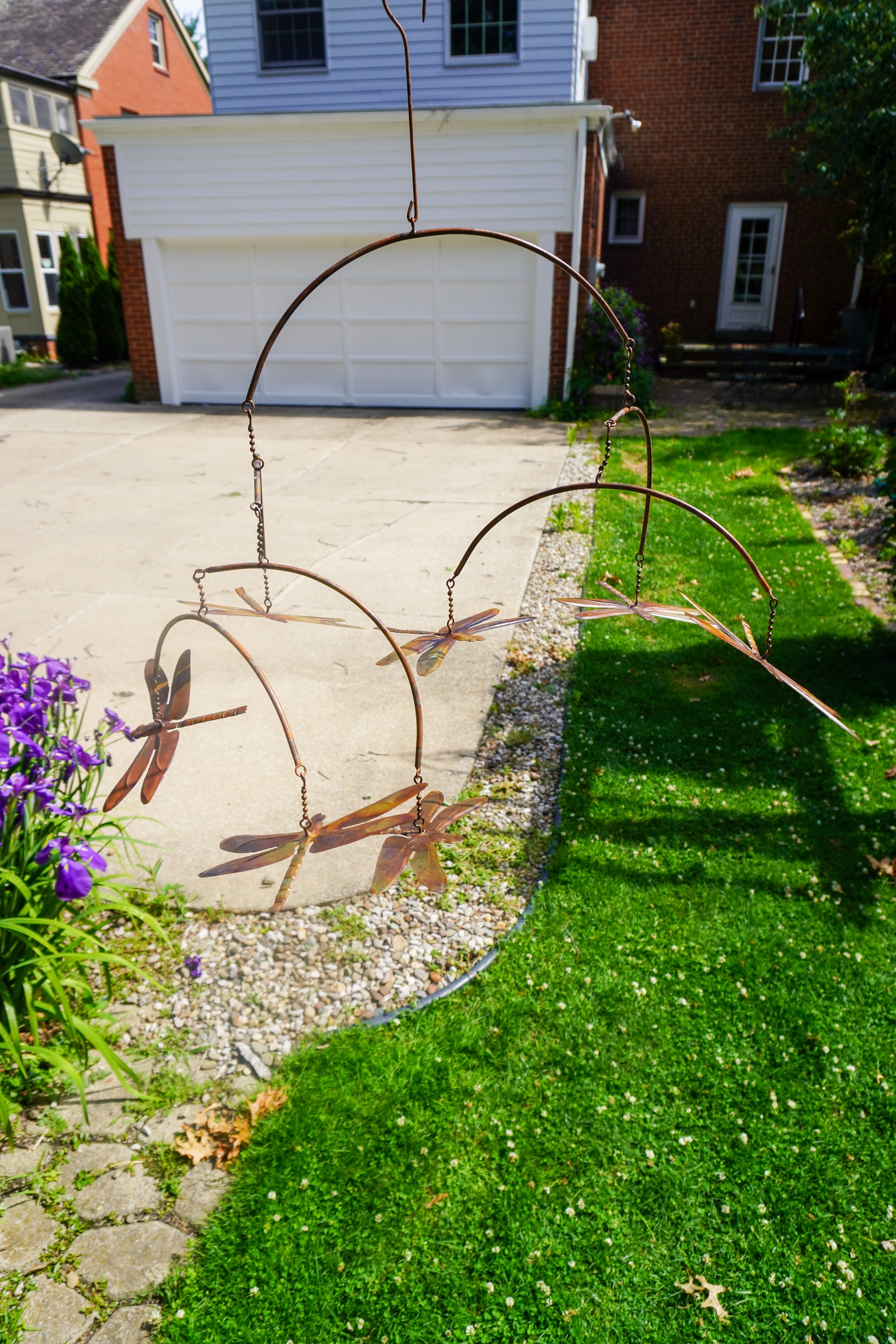 A rusted metal dragonfly garden swing hanging outdoors in a front yard beside a green lawn and concrete driveway, with houses visible in the background.