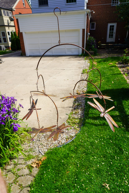 A metal dragonfly garden sculpture with multiple articulated dragonflies hanging from curved wire frames, displayed outdoors on a green lawn next to a concrete driveway and flowers.