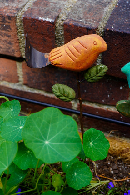 Rusty orange terracotta bird figurine on a metal stake with green cast leaf accents, placed among fern plants