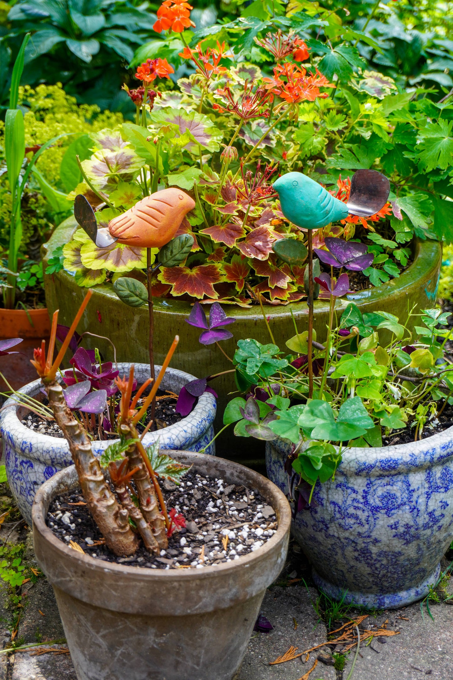 Rusty orange terracotta bird figurine on a metal stake with green cast leaf accents, placed among fern plants