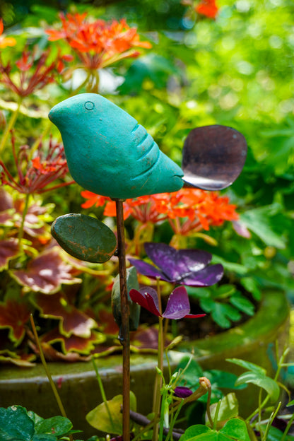 A teal bird-shaped garden stake with a dark leaf-shaped tail, planted in a lush garden with orange flowers and green foliage.