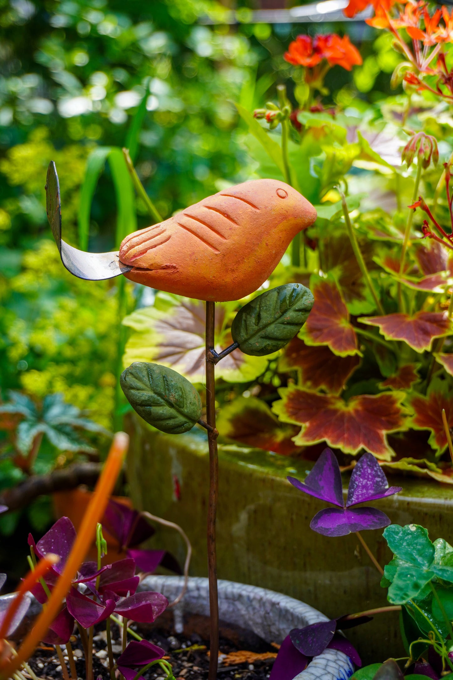 Rusty orange terracotta bird figurine on a metal stake with green cast leaf accents, placed among fern plants