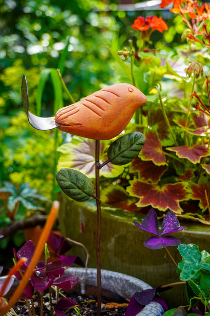 Rusty orange terracotta bird figurine on a metal stake with green cast leaf accents, placed among fern plants