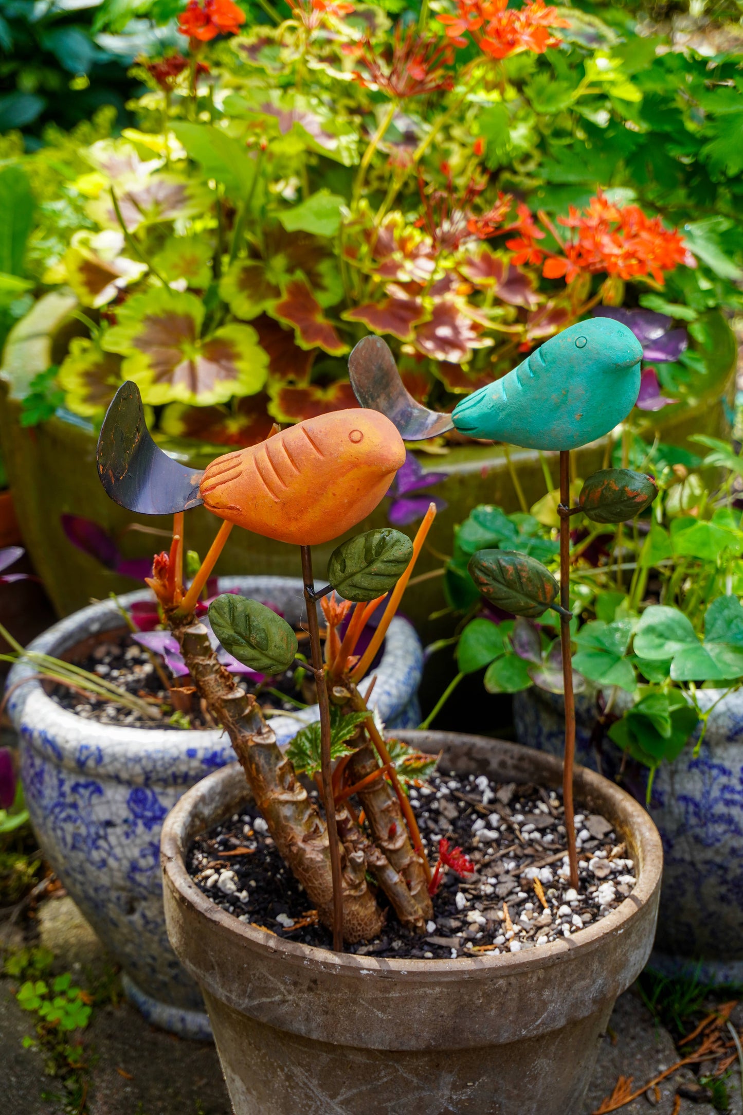 Rusty orange terracotta bird figurine on a metal stake with green cast leaf accents, placed among fern plants