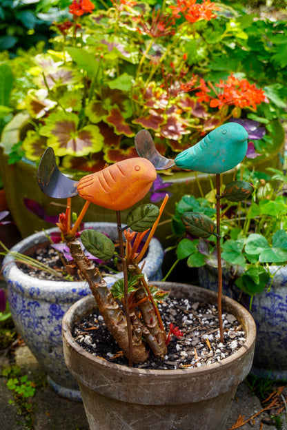 Rusty orange terracotta bird figurine on a metal stake with green cast leaf accents, placed among fern plants