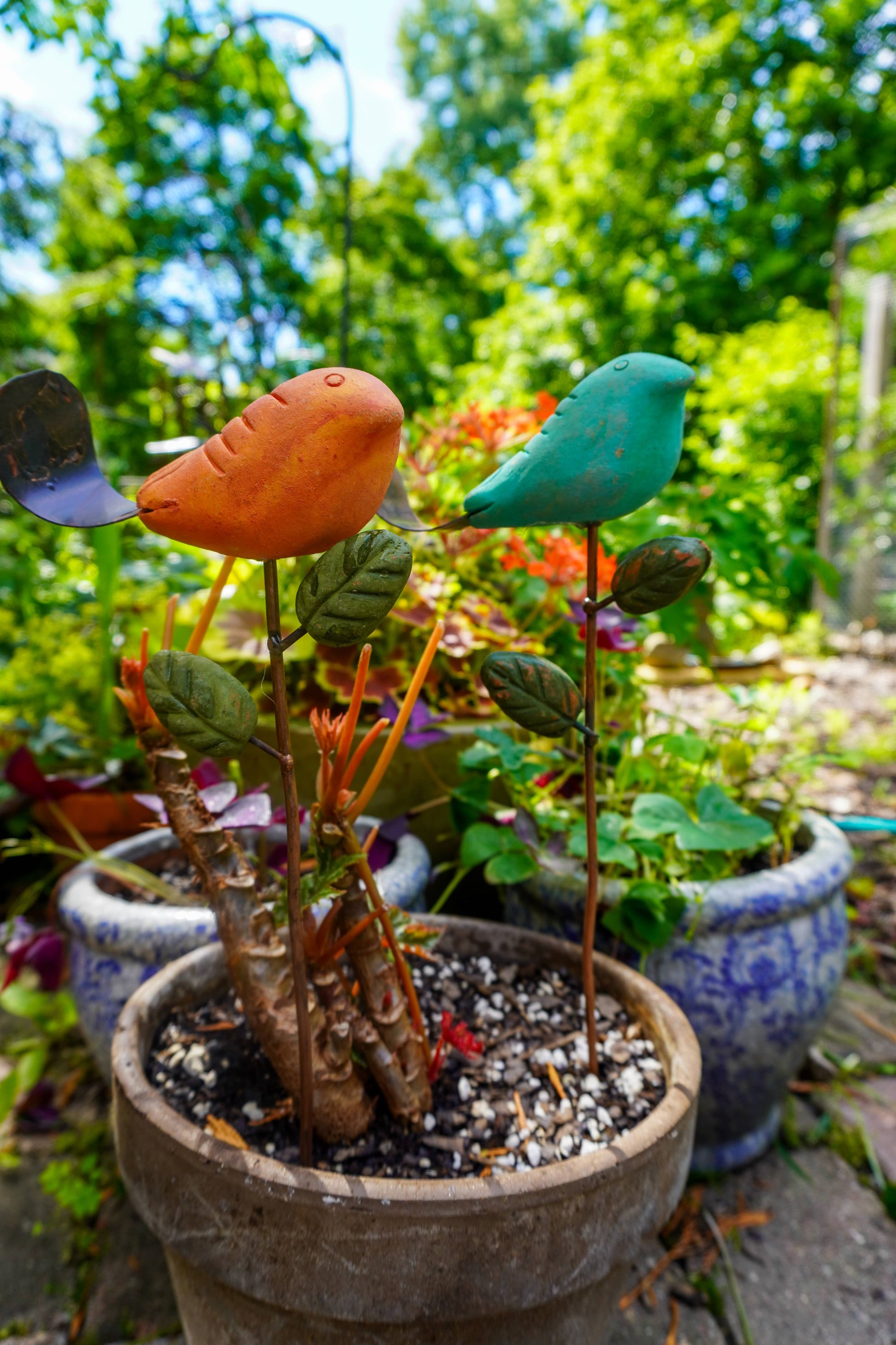 Rusty orange terracotta bird figurine on a metal stake with green cast leaf accents, placed among fern plants