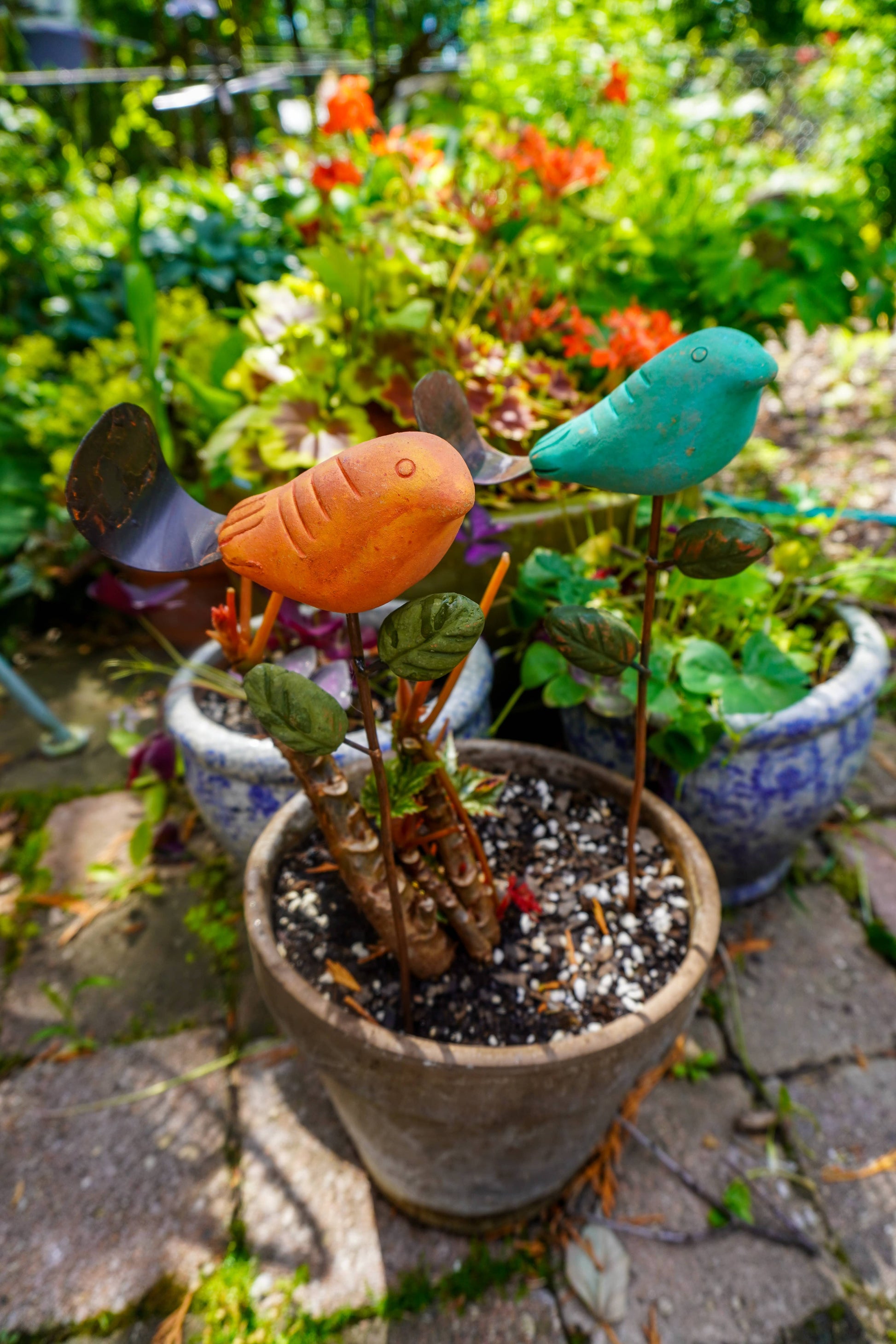 Rusty orange terracotta bird figurine on a metal stake with green cast leaf accents, placed among fern plants