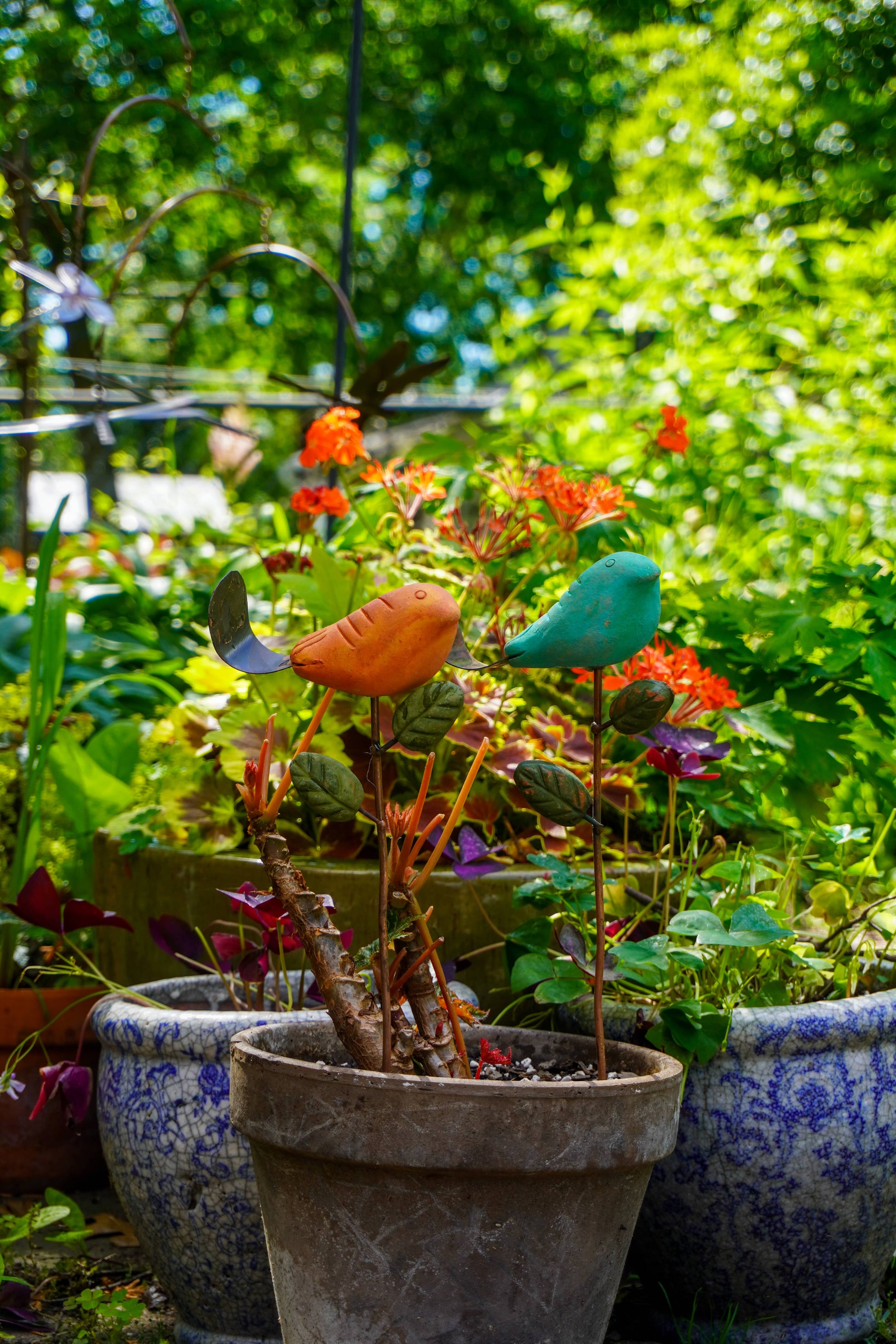Rusty orange terracotta bird figurine on a metal stake with green cast leaf accents, placed among fern plants