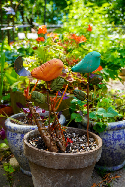 Rusty orange terracotta bird figurine on a metal stake with green cast leaf accents, placed among fern plants