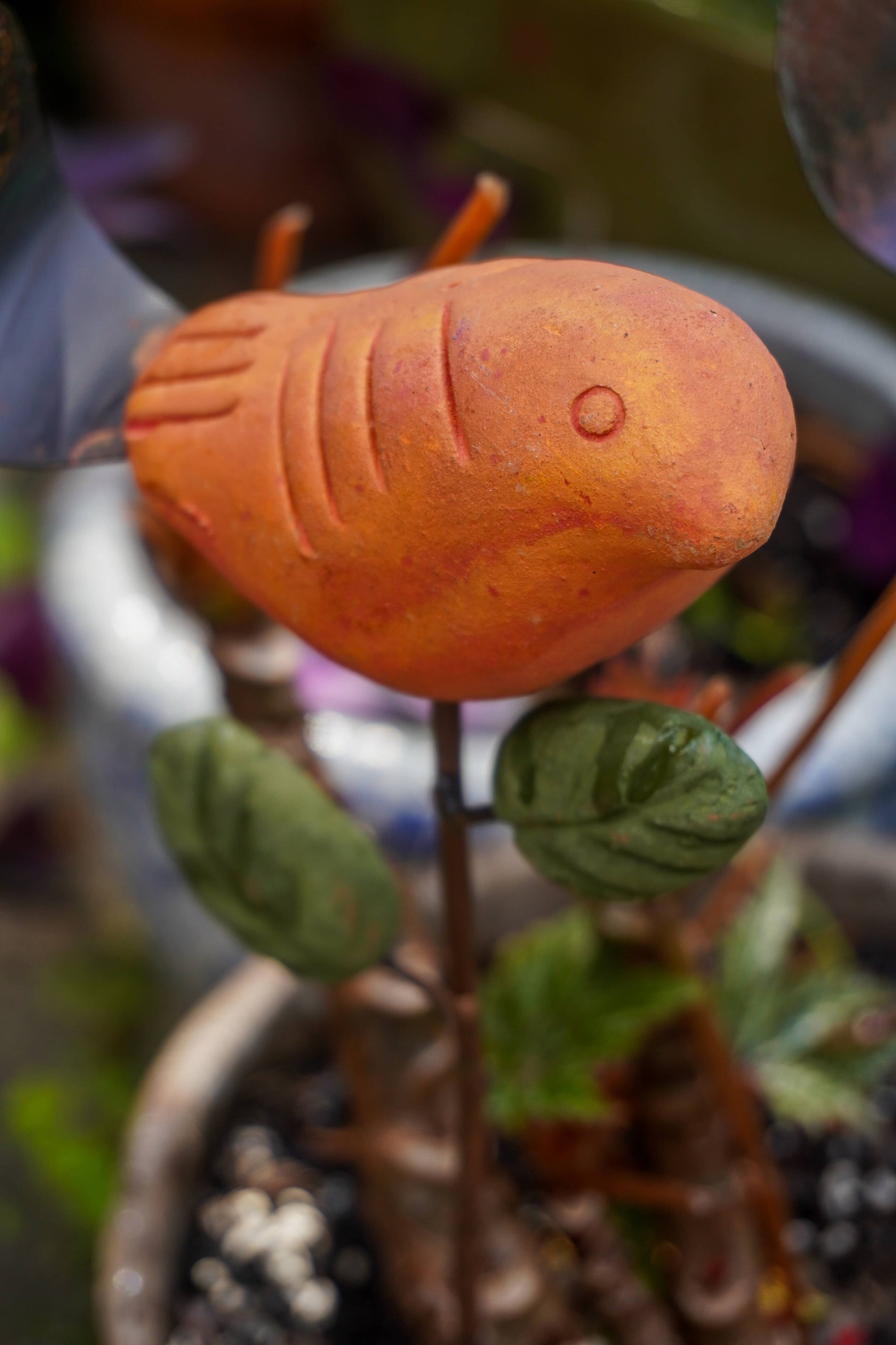 Rusty orange terracotta bird figurine on a metal stake with green cast leaf accents, placed among fern plants