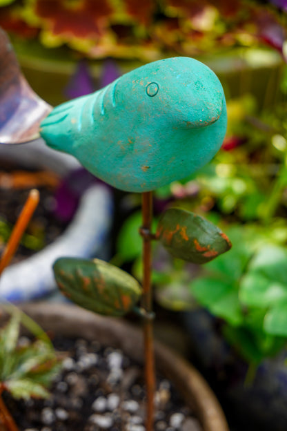 A turquoise painted bird-shaped garden stake with a small leaf accent, photographed in a real garden setting with blurred plants and soil in the background.