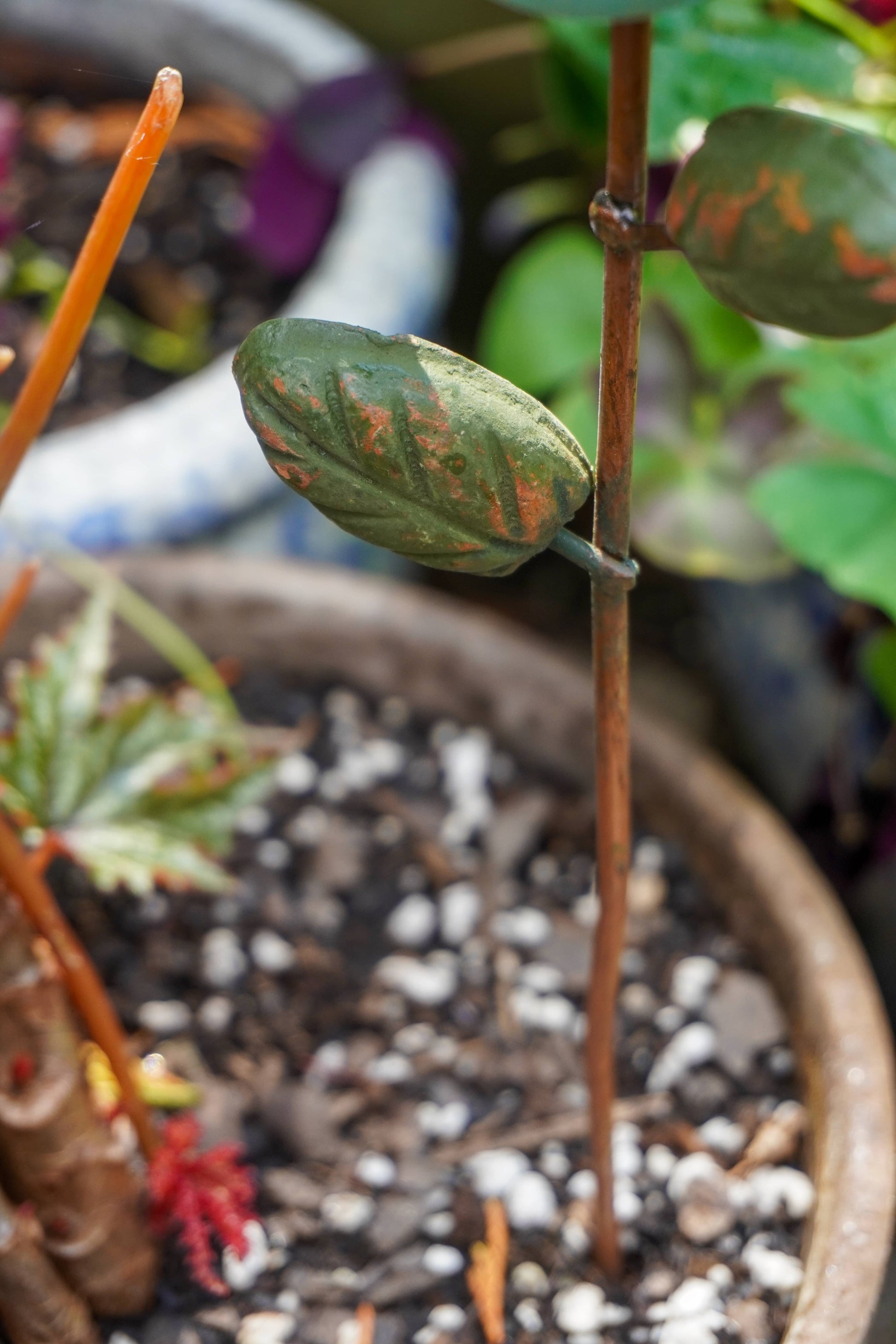 Rusty orange terracotta bird figurine on a metal stake with green cast leaf accents, placed among fern plants