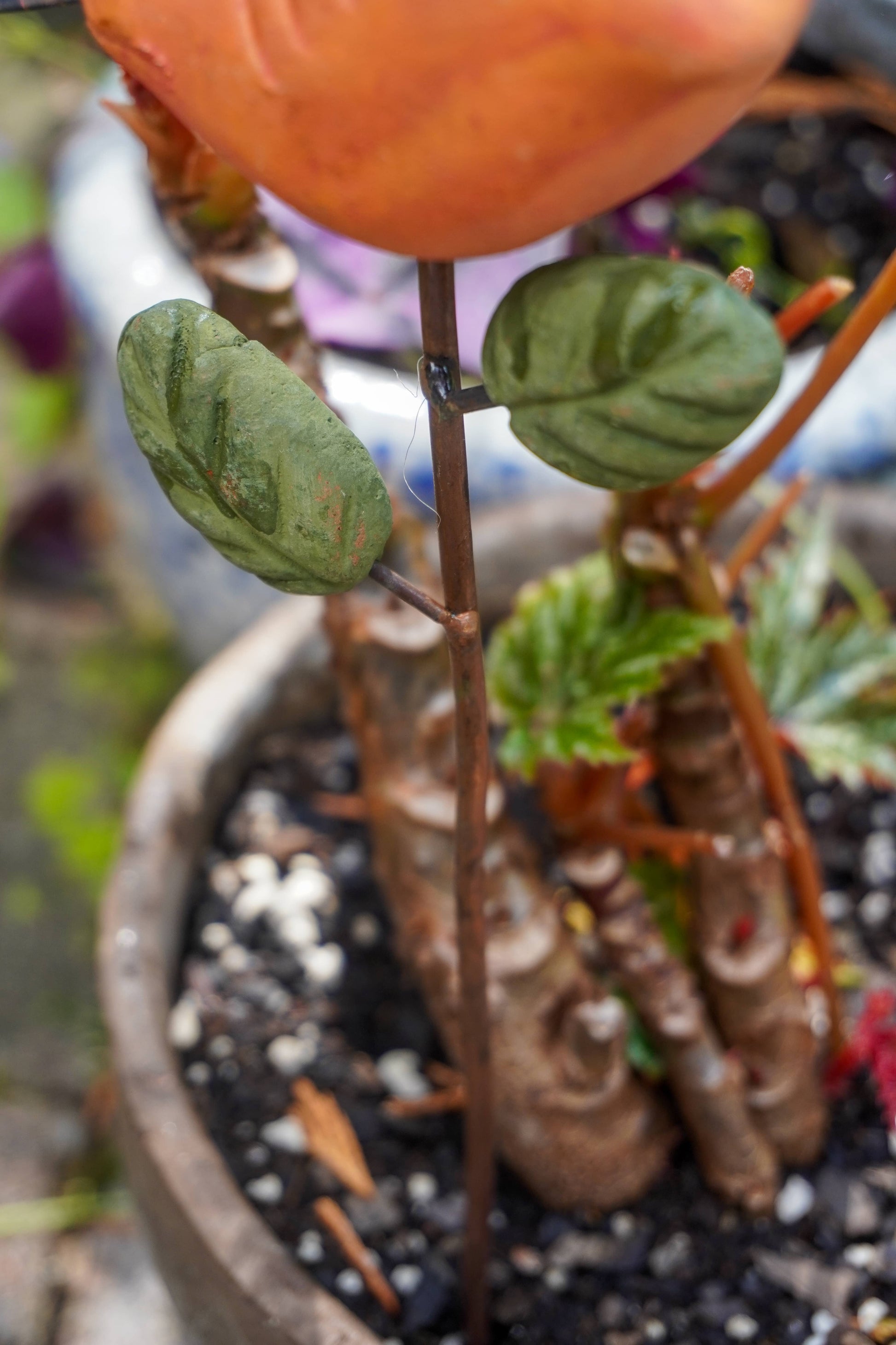 Rusty orange terracotta bird figurine on a metal stake with green cast leaf accents, placed among fern plants