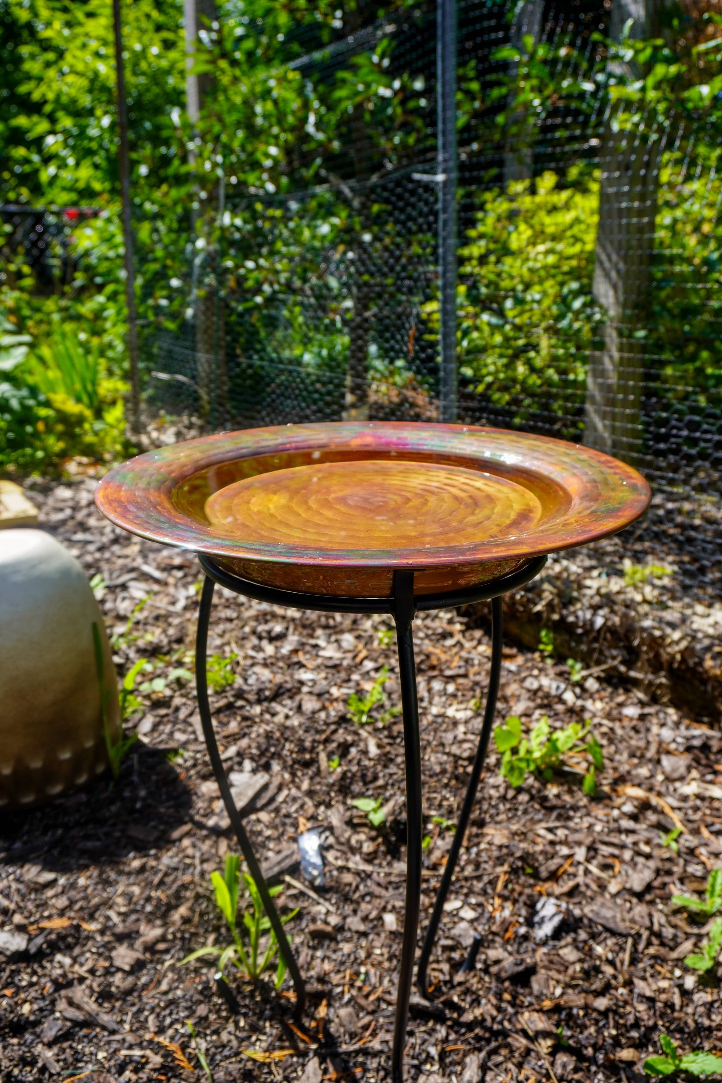 Hammered copper round birdbath with iridescent finish on curved metal stand.