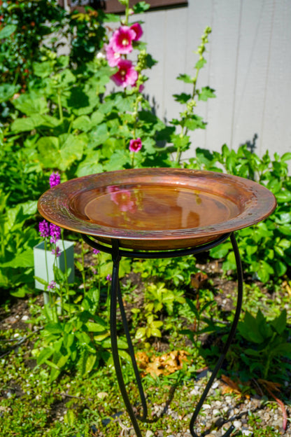 Hammered copper round birdbath with iridescent finish on curved metal stand.