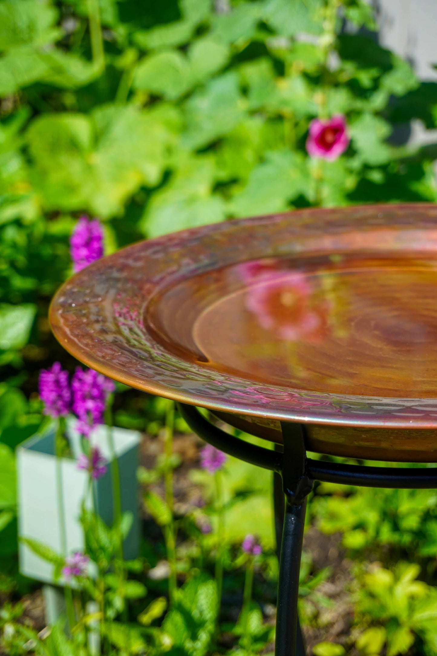Hammered copper round birdbath with iridescent finish on curved metal stand.