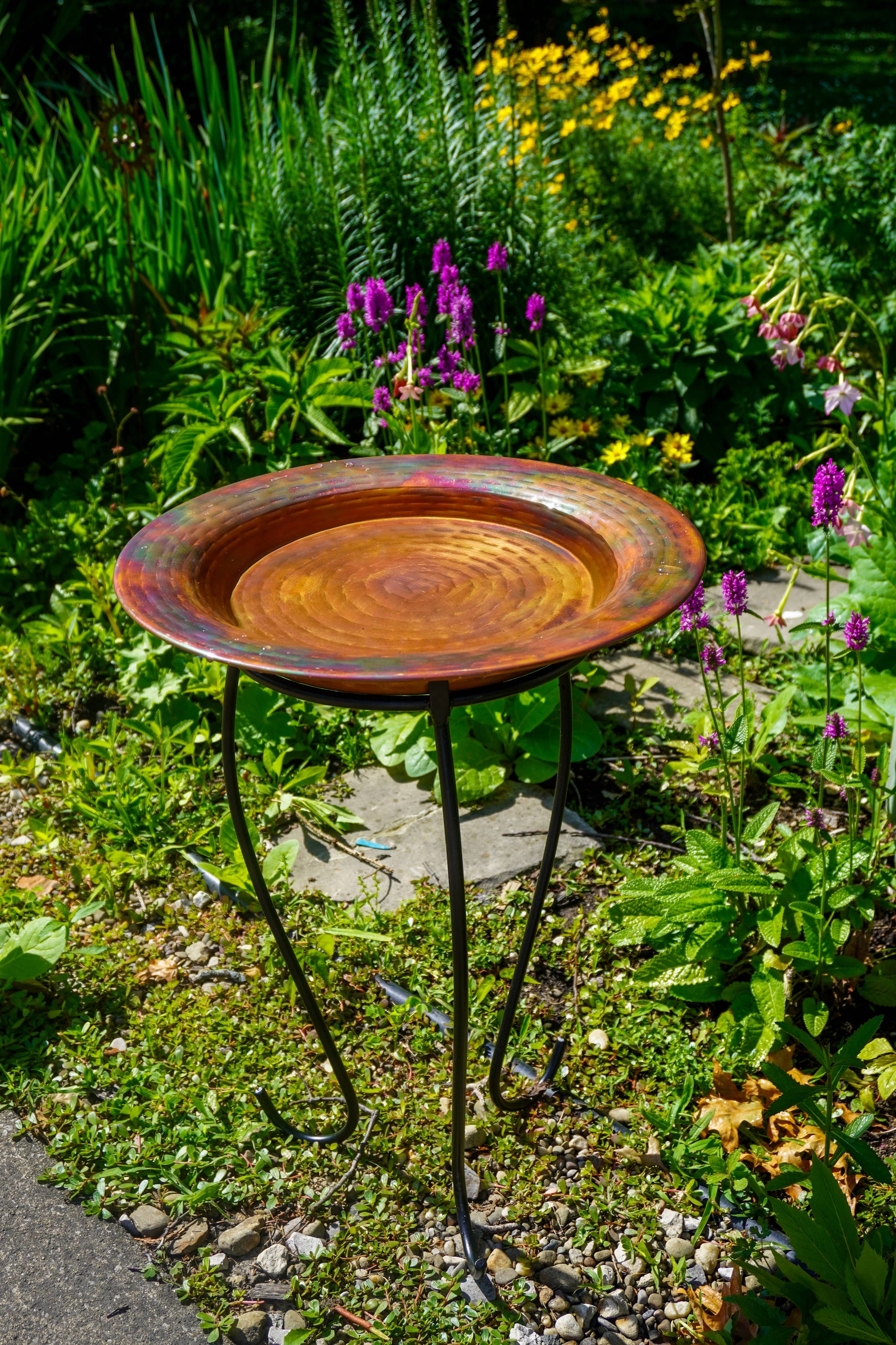 Hammered copper round birdbath with iridescent finish on curved metal stand, holding water with floating red rose petals