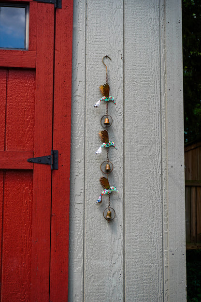 A vertical hanging decorative wall ornament with small gold bells, brown feathers, and colorful beads, displayed against a light gray wooden wall next to a red door.