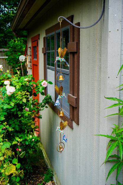 A decorative wind chime with gold-toned birds and blue-and-white patterned accents hangs from a hook outside a house window next to green garden plants.